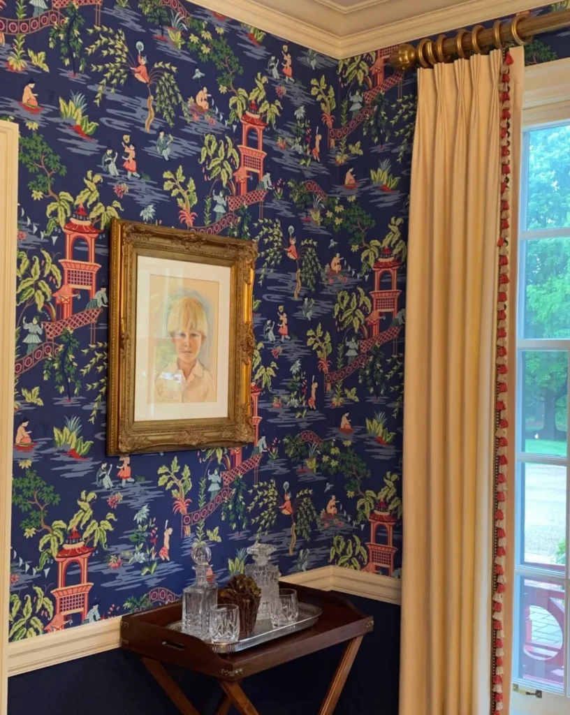 Room corner with blue patterned wallpaper, framed portrait on wall, and a wooden cart holding glassware. Cream curtains are partially drawn over a large window.